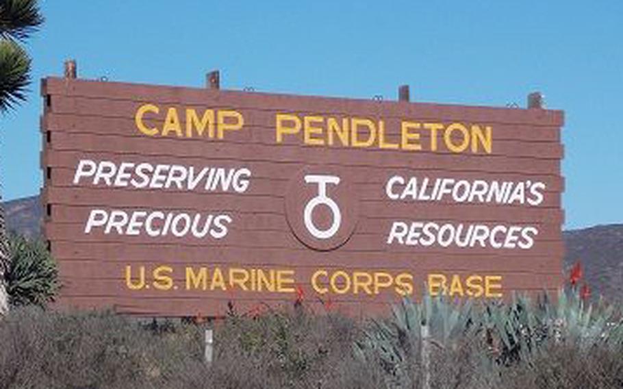 A wooden sign reading “Camp Pendleton U.S. Marine Corps Base” sits on atop a small hill with vegetation around it and blue sky in the background.